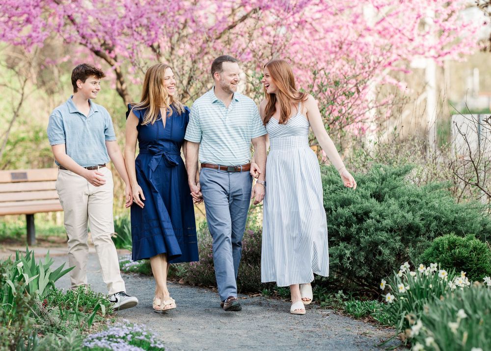 Family of four walking together in a blooming spring garden with pink cherry blossom trees in soft natural light.