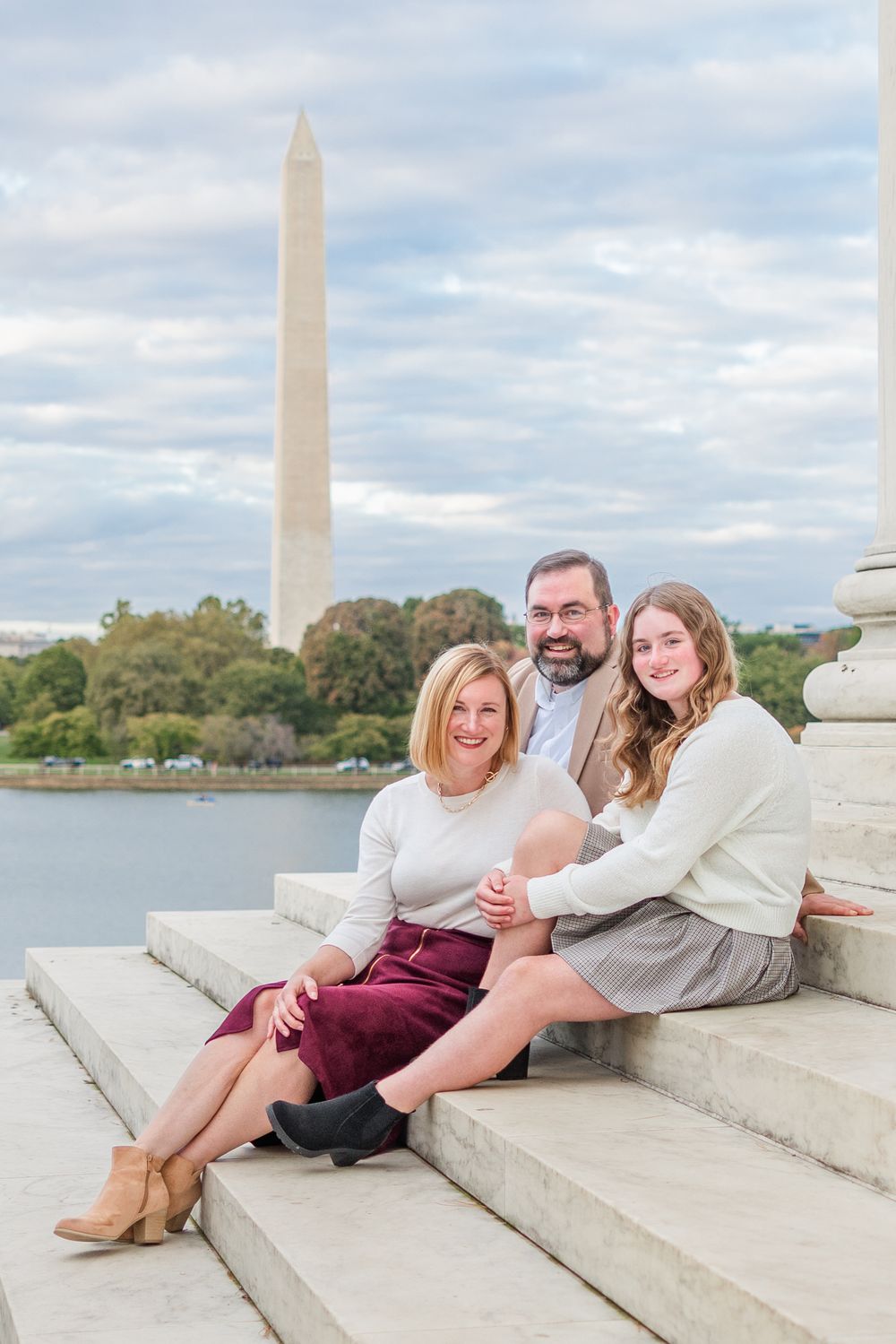 Three people sit together on stone steps with the Washington Monument visible in the background on a cloudy day.