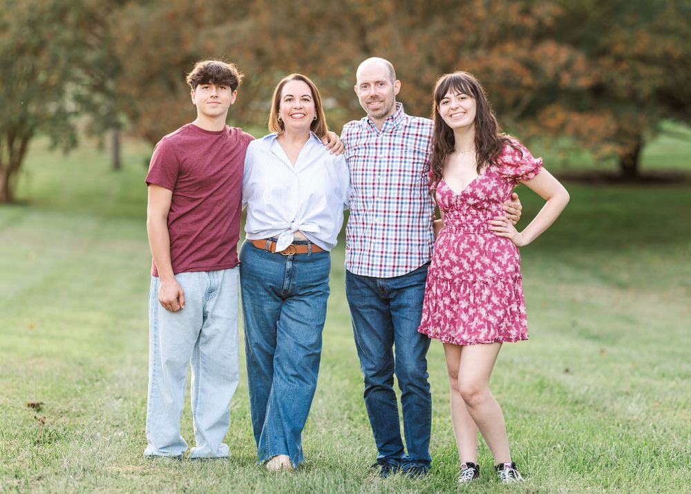 A family of four stands together on grass with trees in the background during a sunny outdoor photo session.