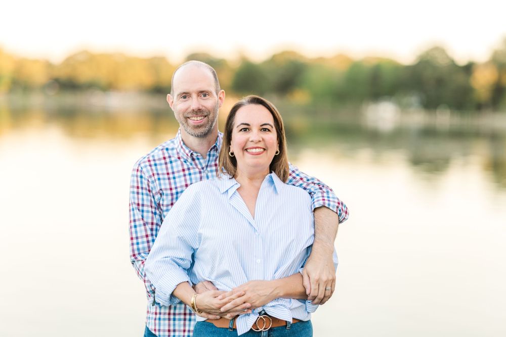 Parents pose sweetly by a serene lake during golden hour at sunset.