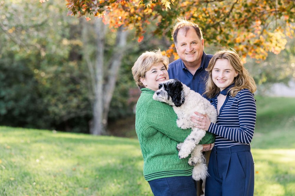 Three people and a small white dog pose together under autumn trees in a park setting during golden hour.