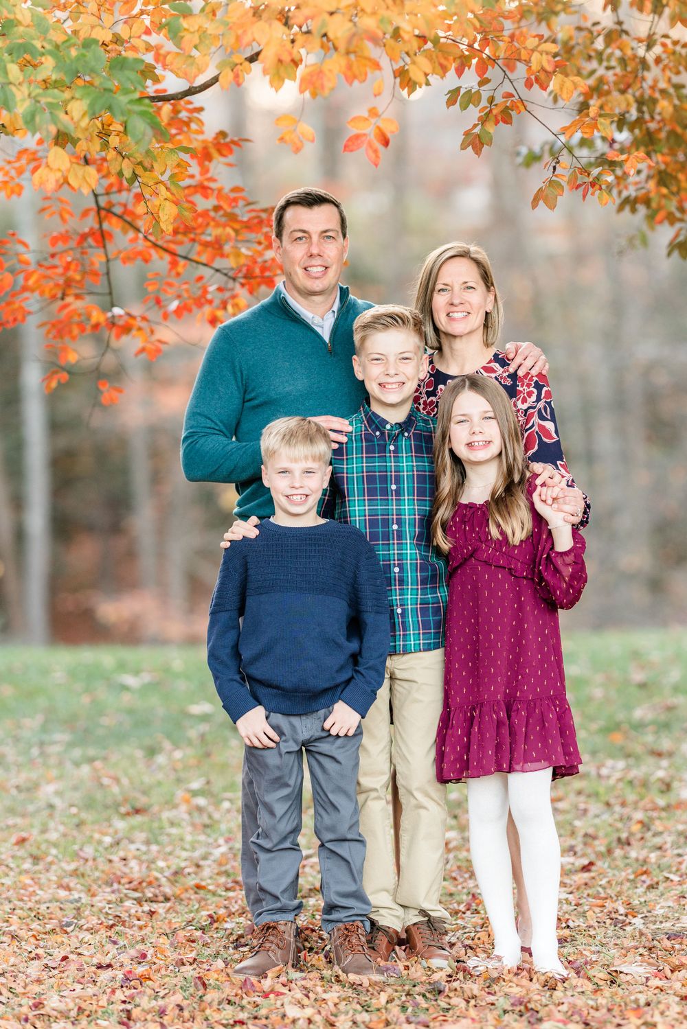 A family of five poses together under vibrant orange autumn leaves with a forest-like setting in the background.