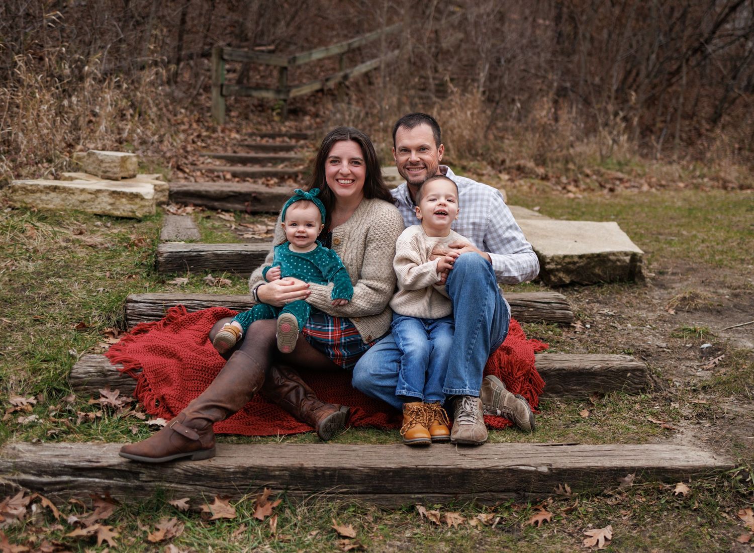 A family sits together on rustic stone steps outdoors with a red blanket, surrounded by autumn woods.