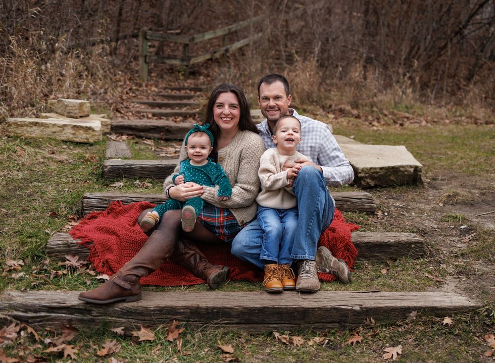 A family sits together on rustic stone steps outdoors with a red blanket, surrounded by autumn woods.