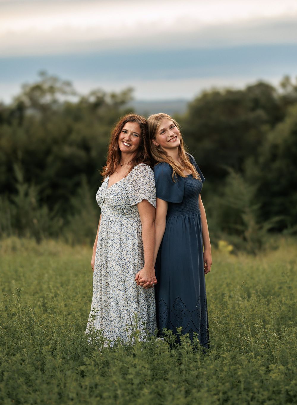 Two people in floral and navy dresses stand together in a sunlit meadow during golden hour.