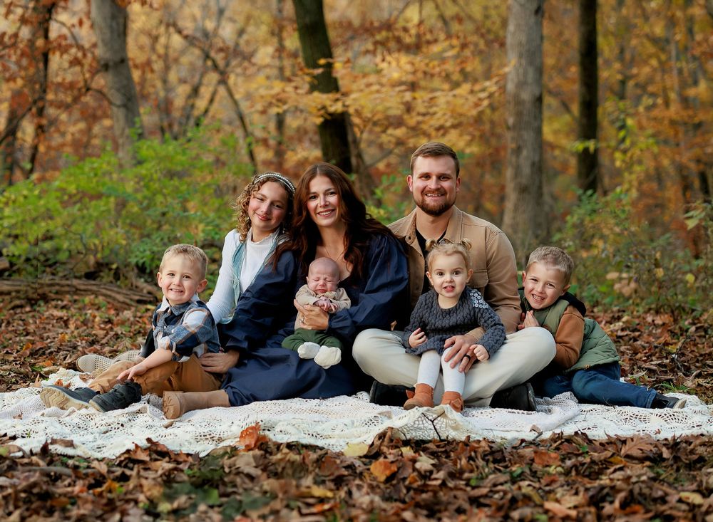 A group relaxes together on a white blanket surrounded by fallen autumn leaves in the woods.