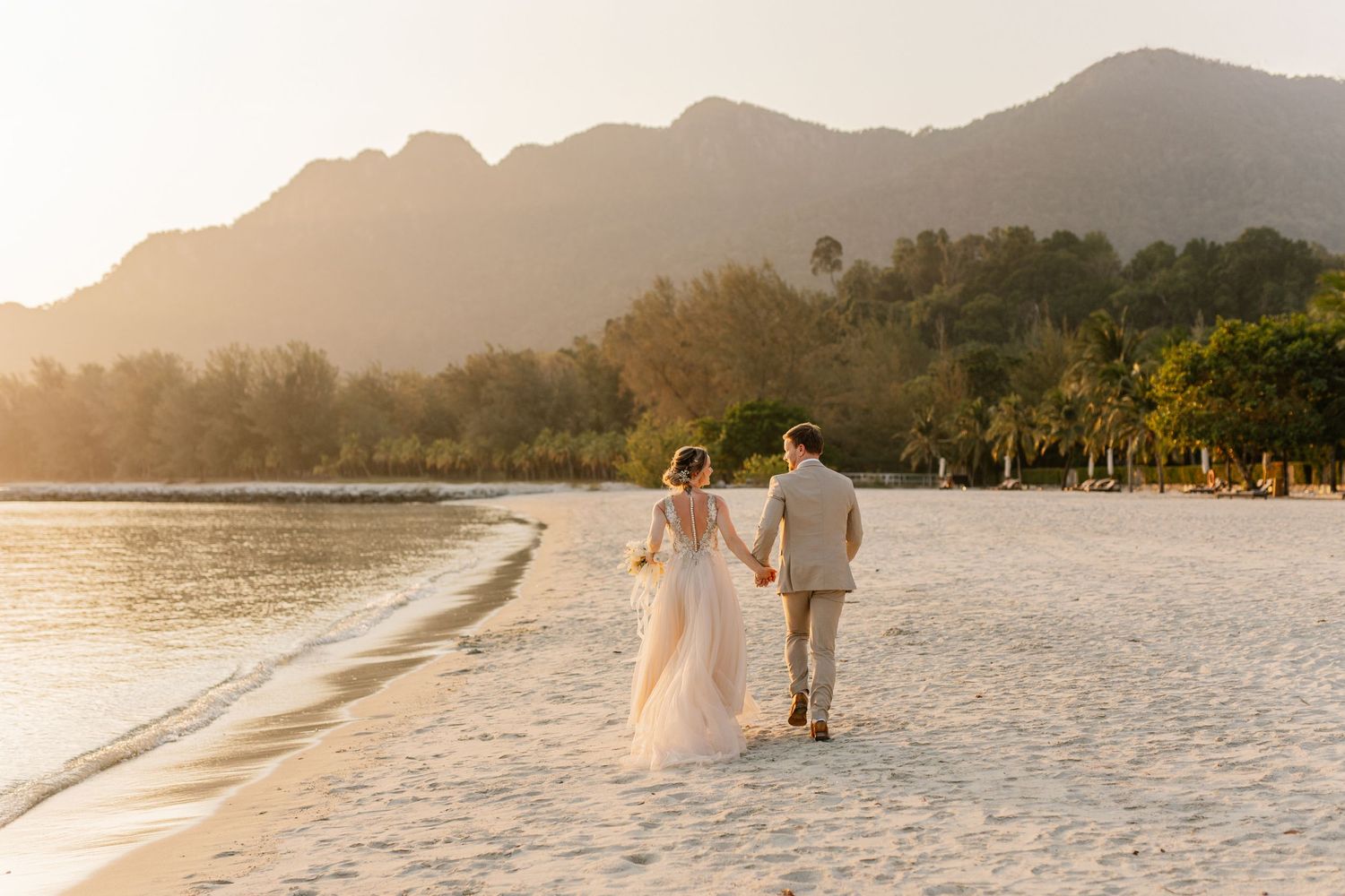 A couple walks hand in hand along a tropical beach at sunset with mountains and palm trees in the background.