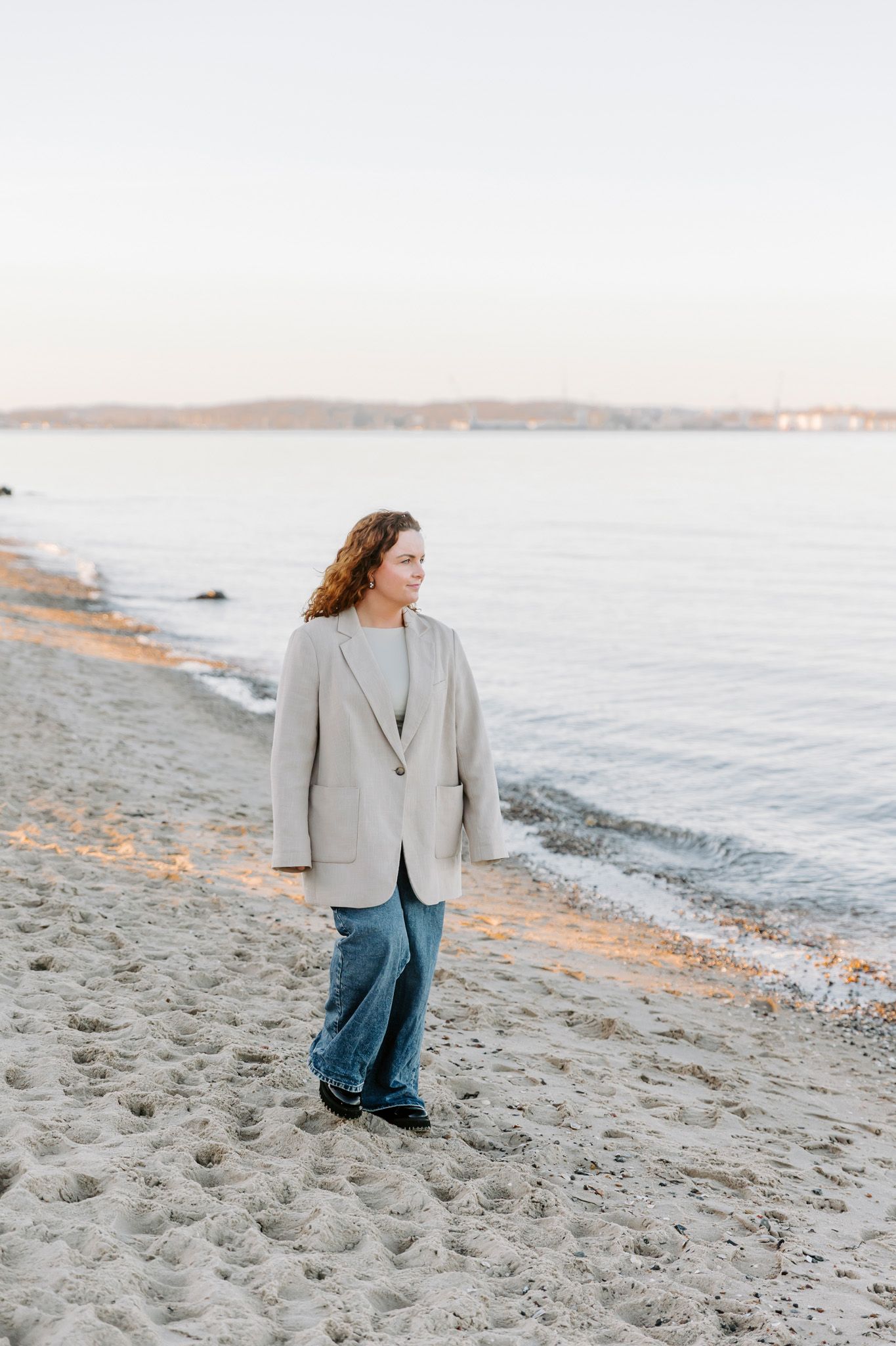 En person i en grå sweater og jeans går langs en sandstrand ved solnedgang, mens bølgerne ruller ind. Branding fotograf Fyn.
