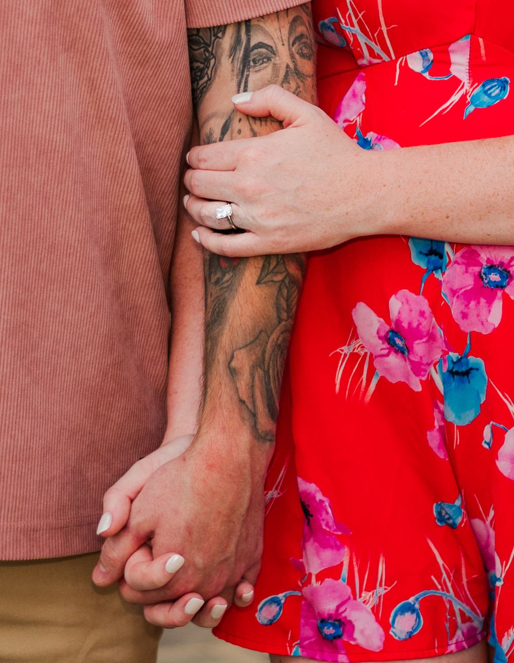 Close-up of hands with engagement ring and detailed arm tattoo against red floral dress.