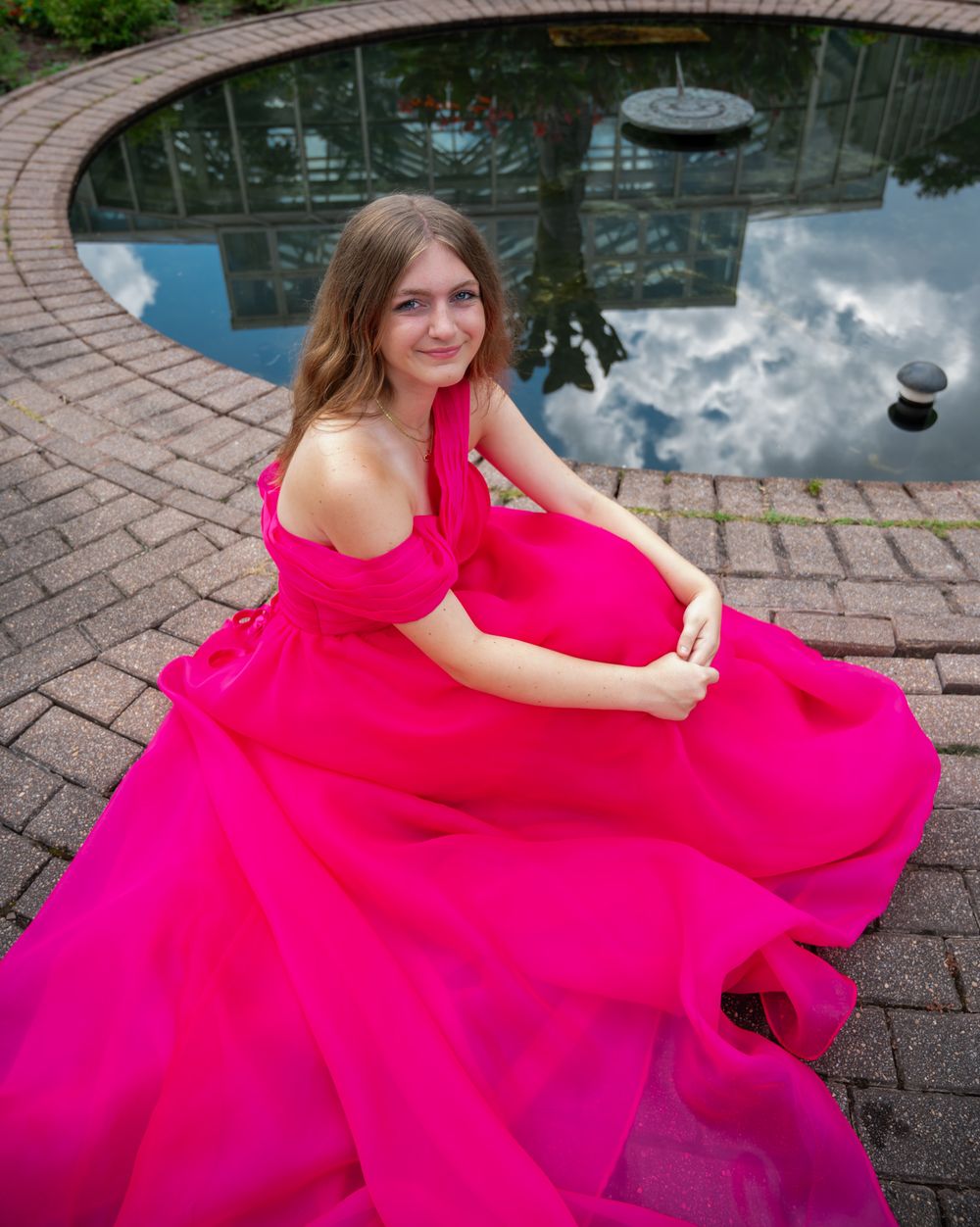 Flowing hot pink formal gown spreads dramatically around model seated by reflecting pool.