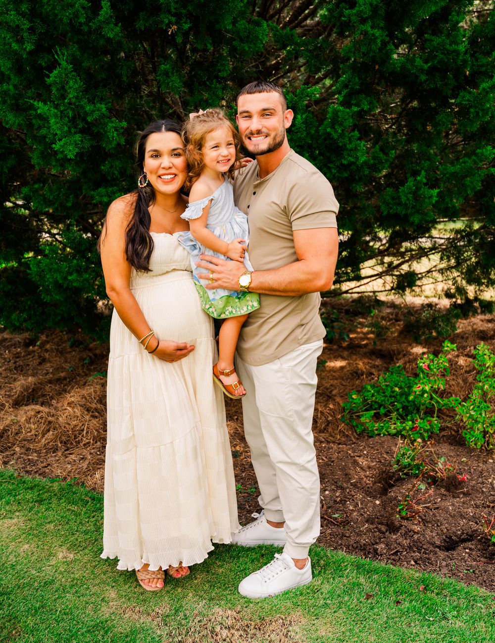 A family poses outdoors against greenery with the mother wearing a white dress and father in casual attire.