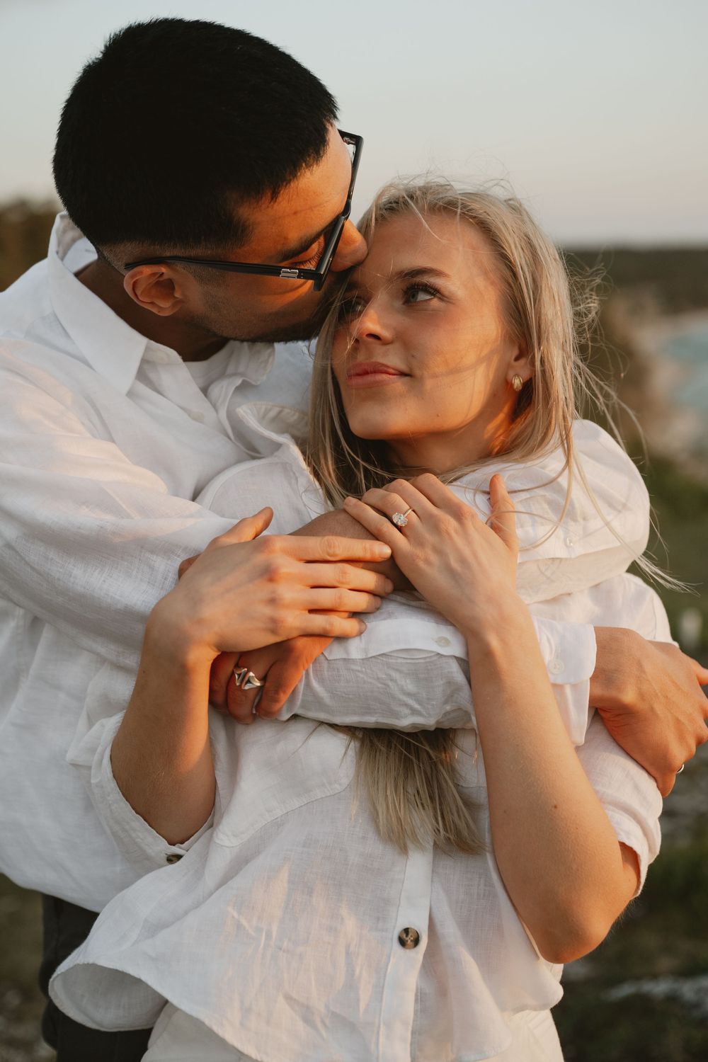 Couple in white linen shirts embracing at sunset, woman smiling up at man wearing glasses in romantic outdoor portrait.