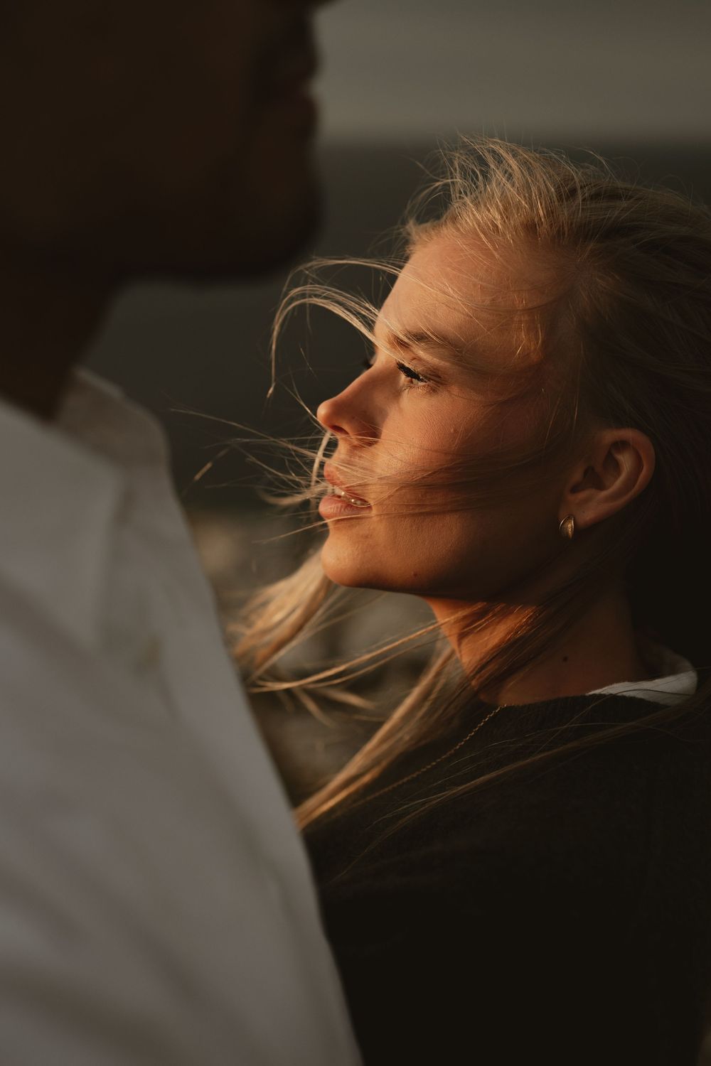 A woman with windswept blonde hair gazes up in golden sunset light, standing close to a man in a white shirt.