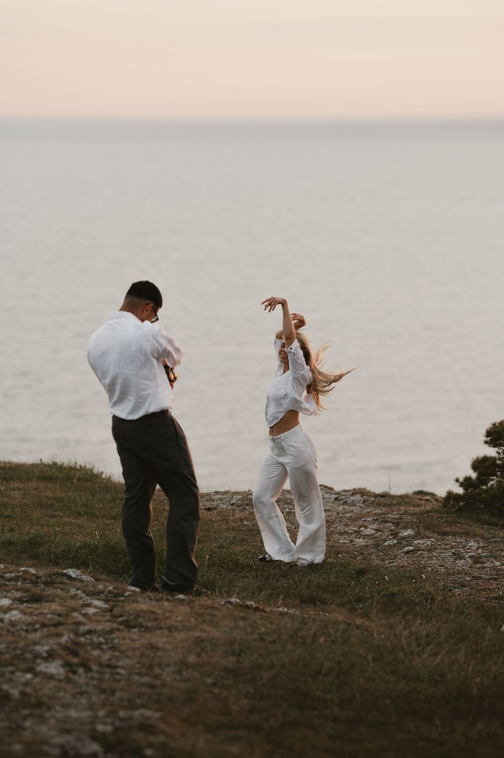 Couple dancing together on a grassy cliff overlooking the ocean at sunset, both wearing white outfits.