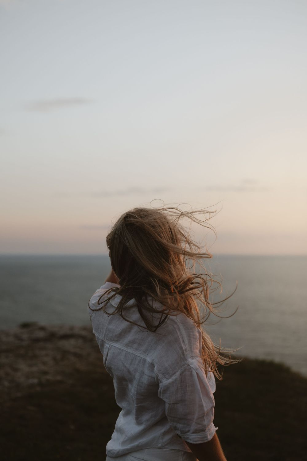 Woman with windswept blonde hair in white shirt gazing at ocean during sunset from coastal cliffside.