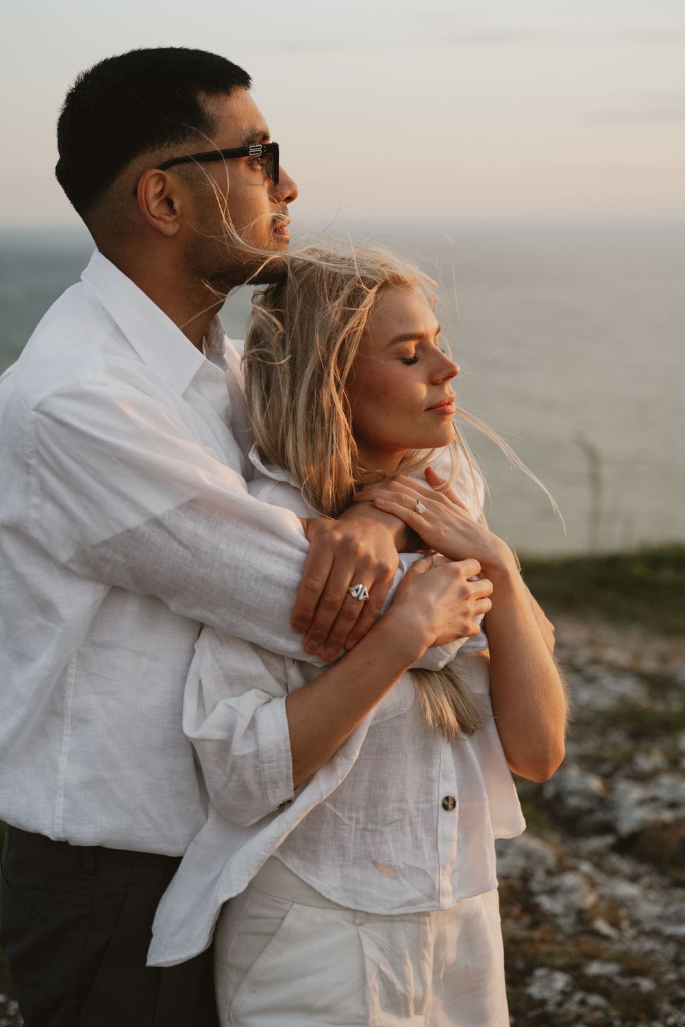 Couple in white linen embracing at sunset by the sea, woman with engagement ring, eyes closed, romantic outdoor portrait.