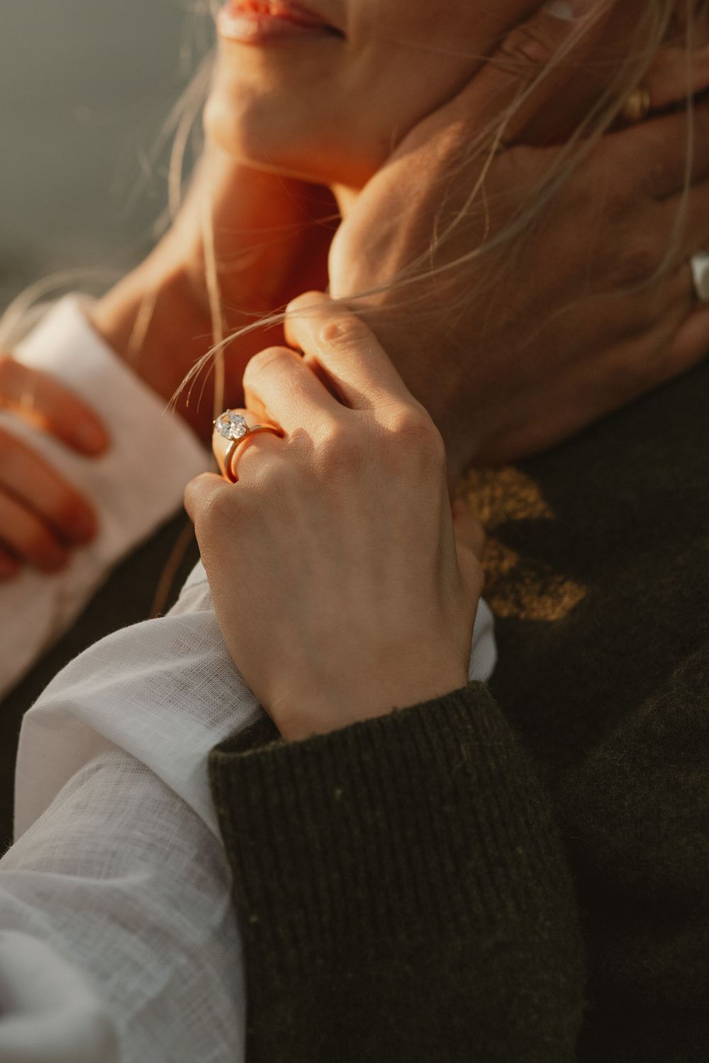 Close-up of a woman's hand wearing a gold diamond engagement ring, touching her hair in warm golden sunlight.