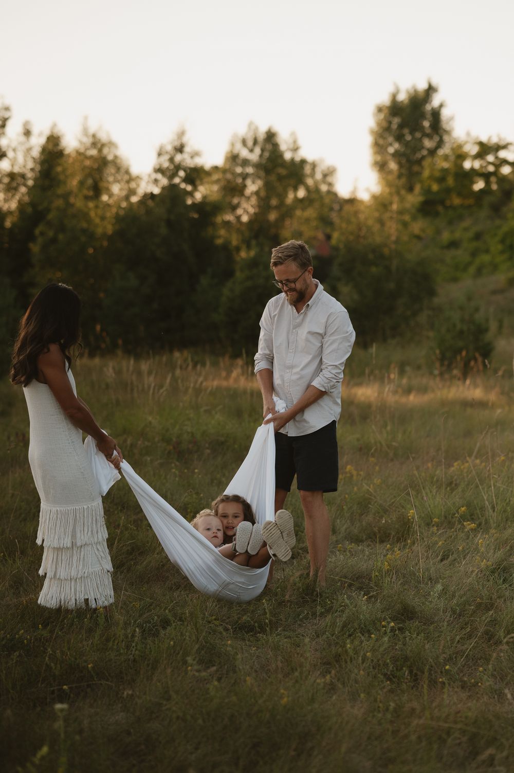 A couple cradles their newborn in a white fabric hammock outdoors in a golden meadow at sunset.