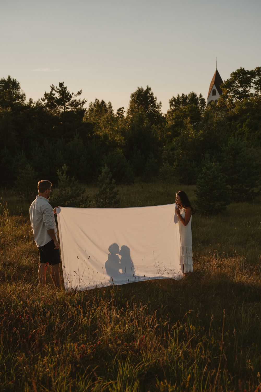 Two people hold a white sheet displaying kissing silhouettes in a field at sunset near a small chapel.