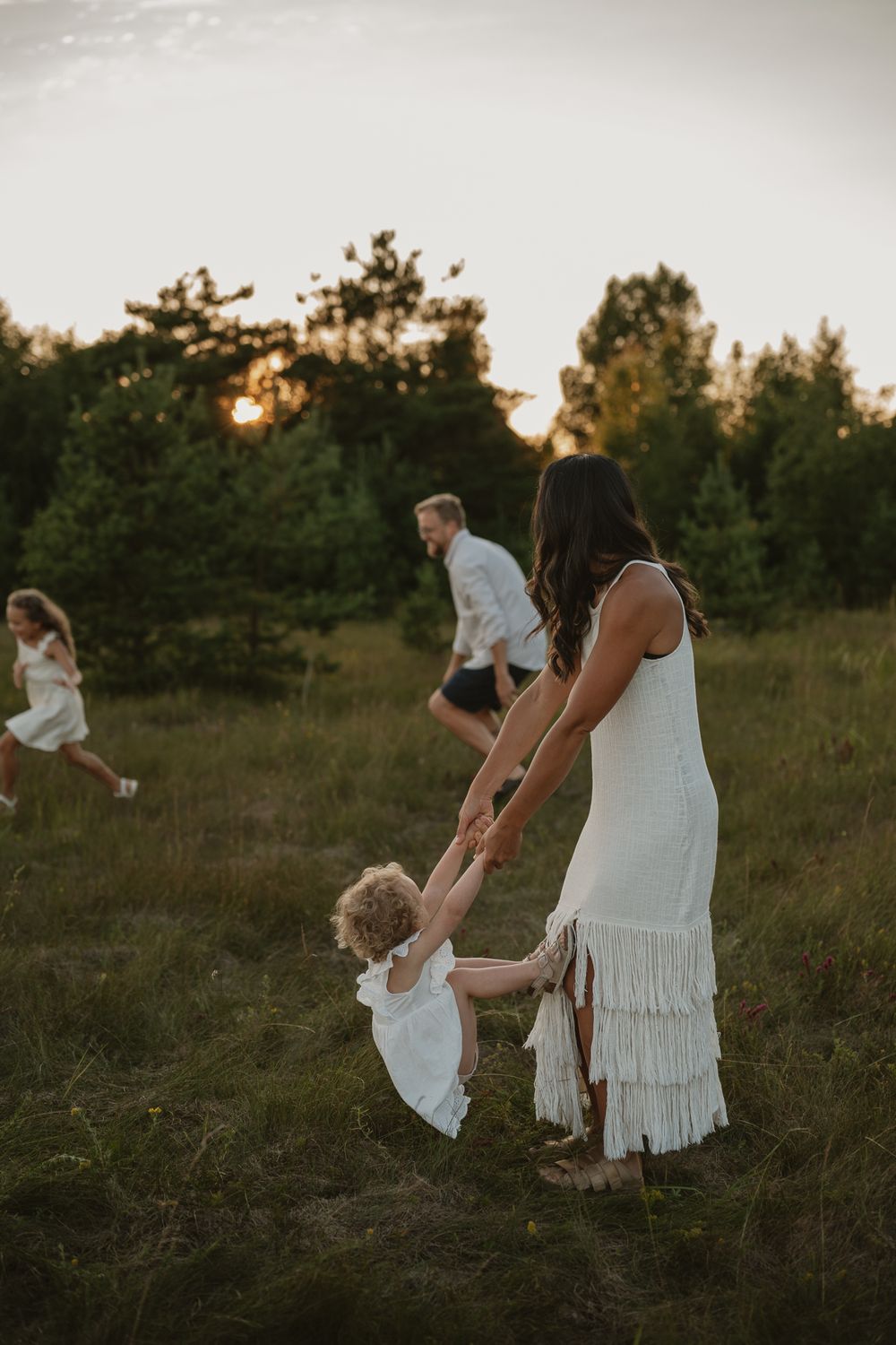 Family in white outfits playing in a green meadow at golden hour sunset, mother holding toddler's hands.