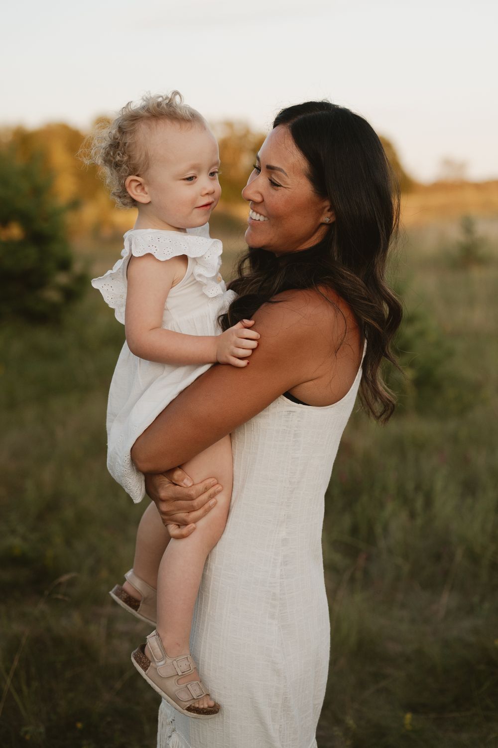 A smiling mother holds her toddler daughter in a sunlit meadow, both wearing white outfits during golden hour.