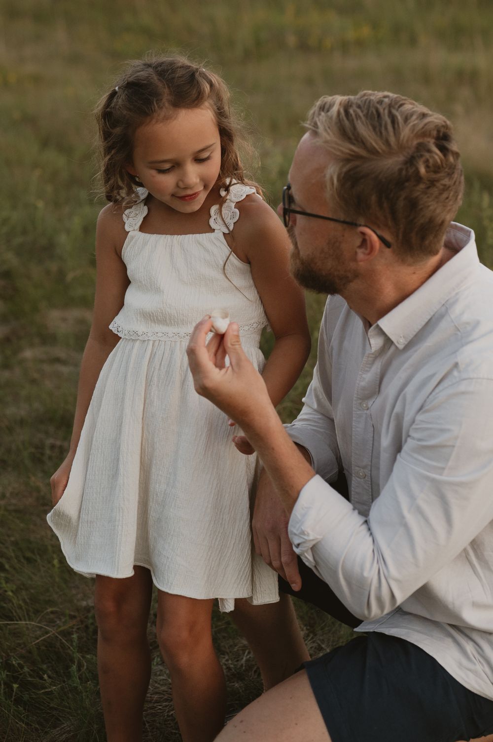 A father kneels in a field, handing a small flower to his young daughter in a white dress.