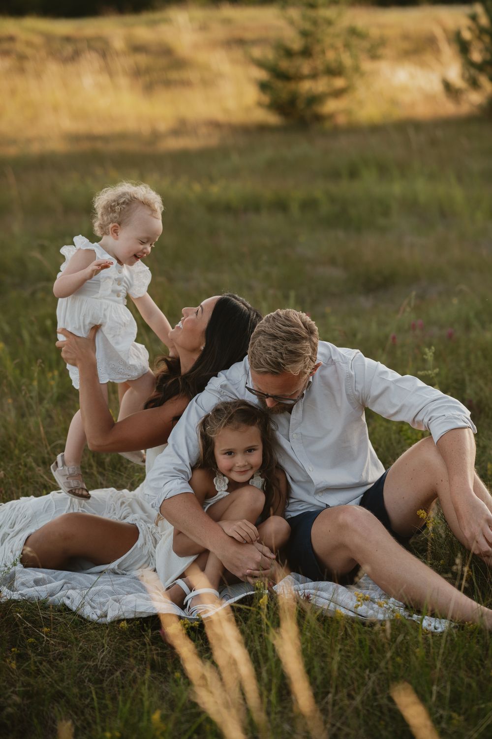 Family of four enjoying a golden hour picnic in a meadow, parents kissing while holding two young children.