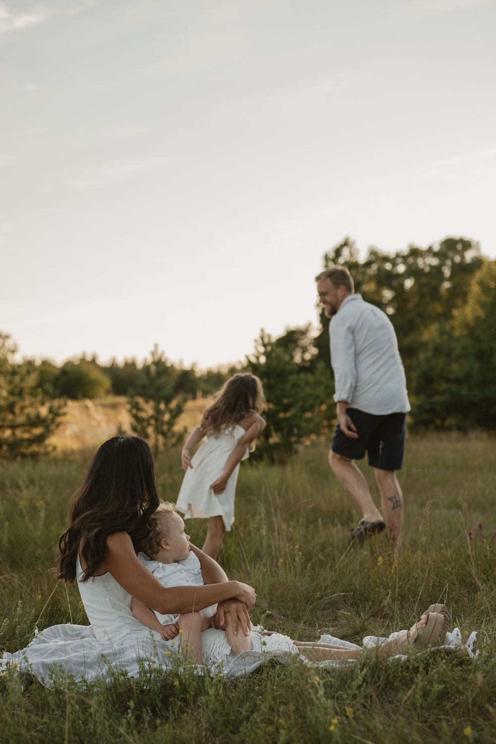 Family enjoying a golden hour picnic in a green meadow, mother holding baby while father plays with young daughter.