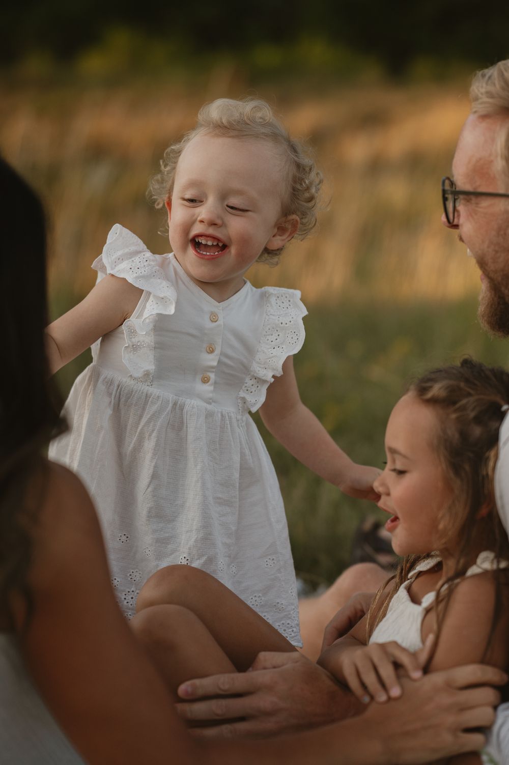 A joyful toddler in a white dress laughs while being held by family members outdoors in a sunny field.