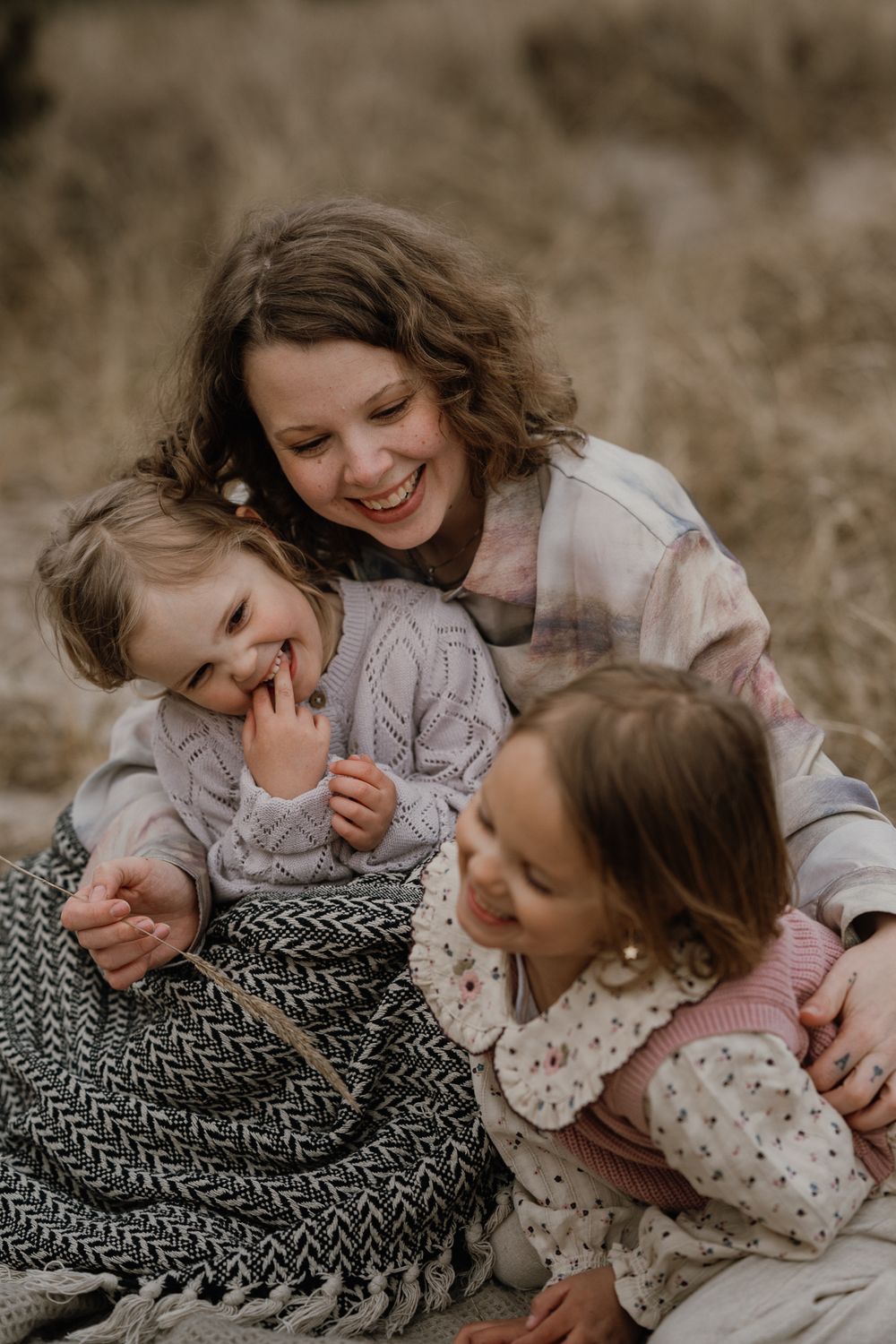 A smiling mother sits outdoors on a blanket, laughing and cuddling her two young daughters in cozy knit sweaters.