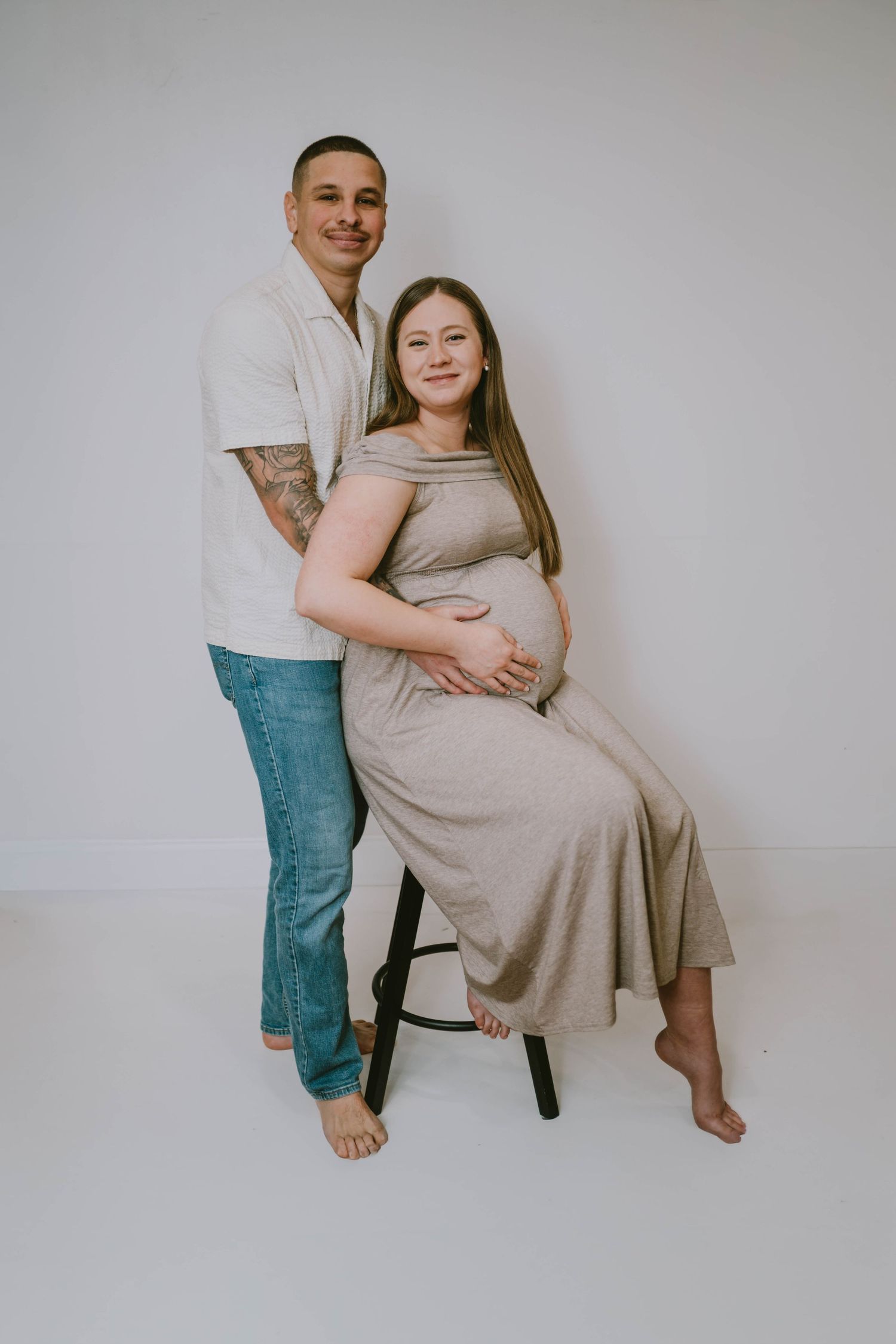 Intimate maternity session with couple sitting together on chair against white backdrop.