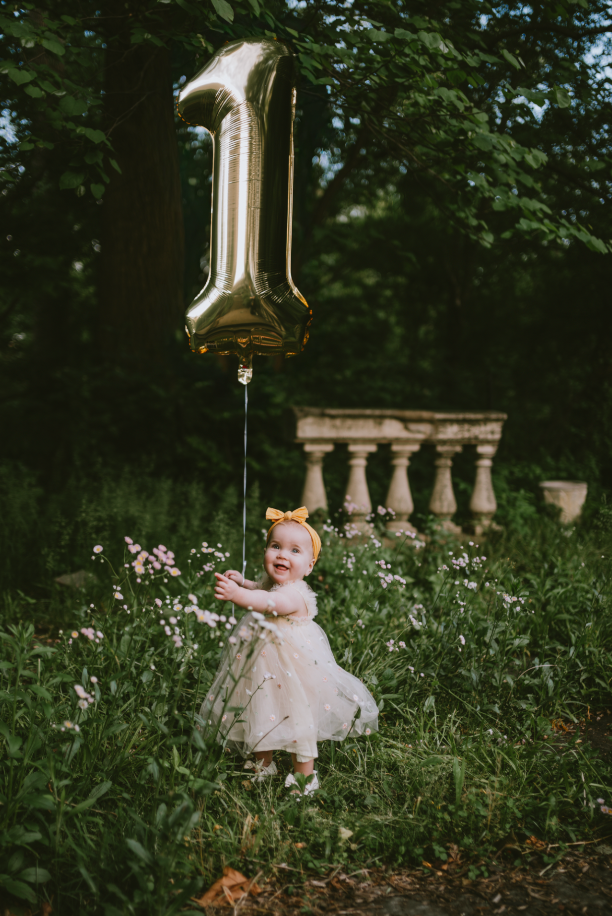 A baby girl in a white dress holds a gold number one balloon in a lush garden, celebrating her first birthday.