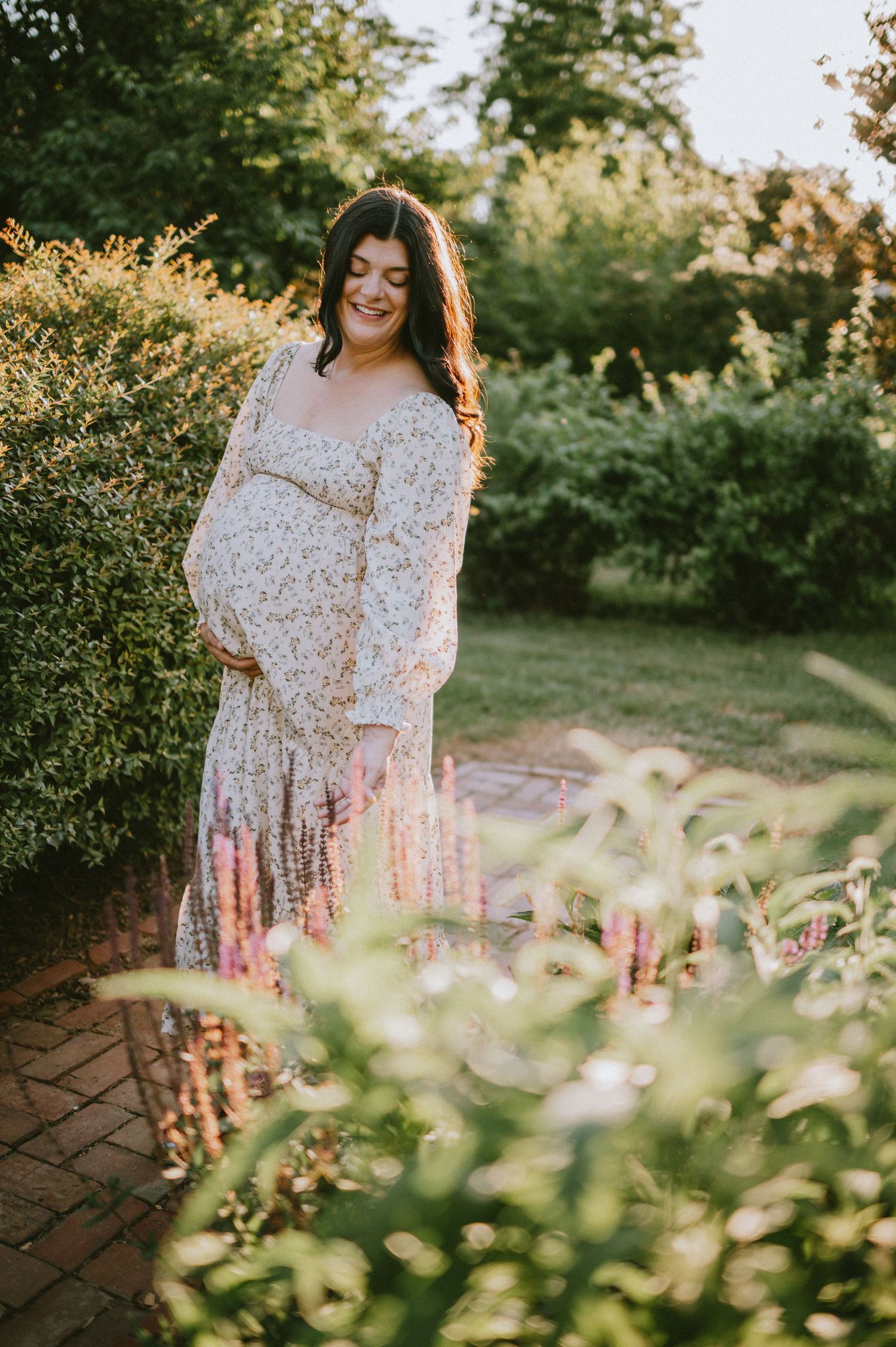 Pregnant woman in floral dress cradling baby bump in a lush garden during golden hour.