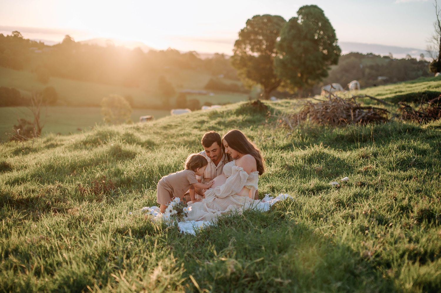 An expectant mother in a white dress stands in golden sunset light.