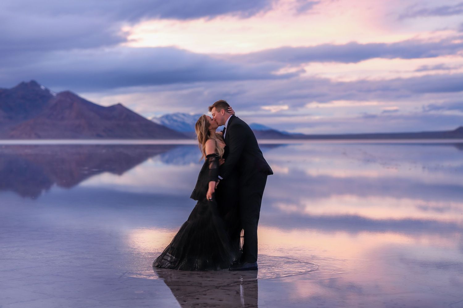 A silhouetted figure reflected in calm waters at sunset with mountains in the background.