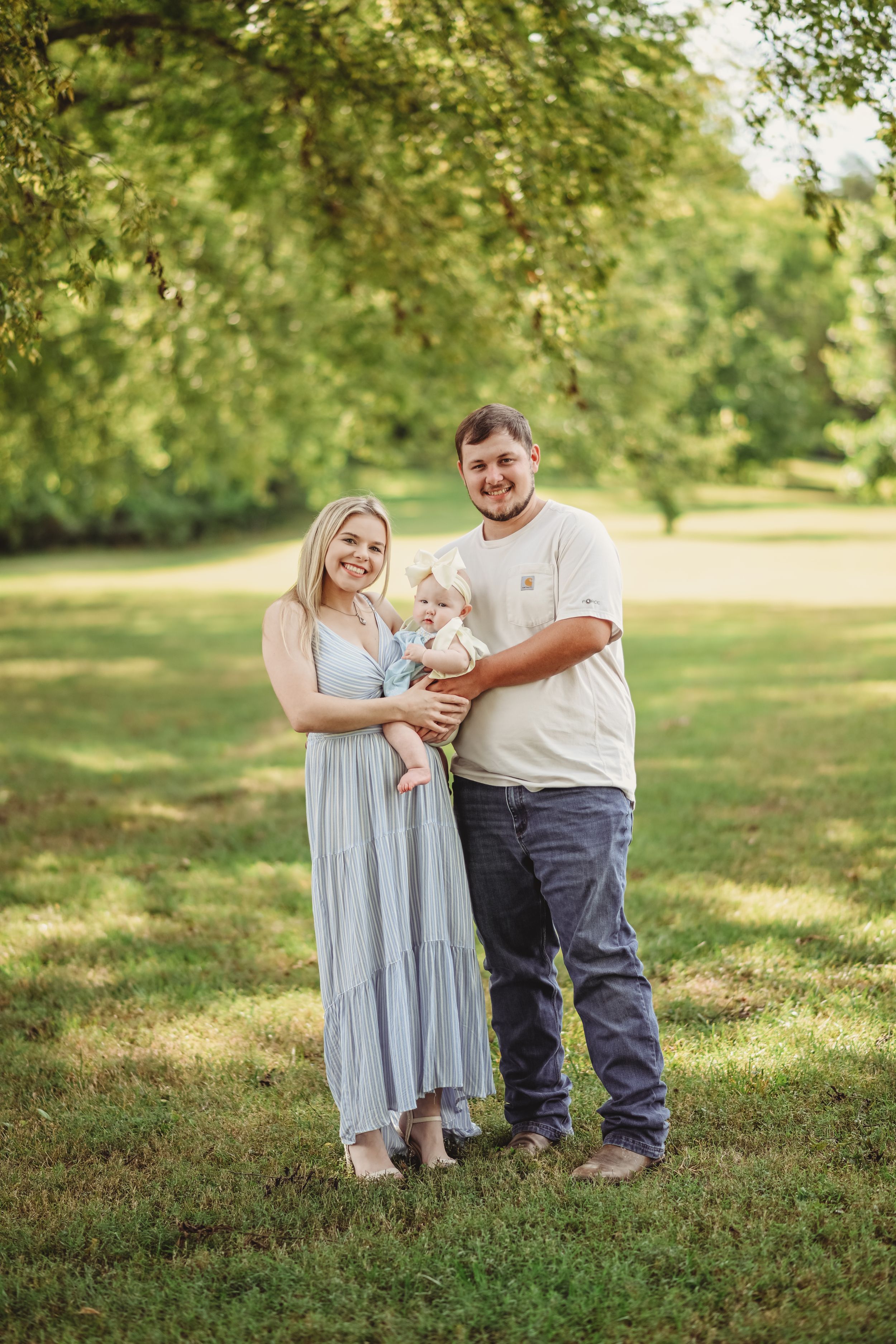 A family poses together on grassy lawn under trees during golden hour outdoor photoshoot.