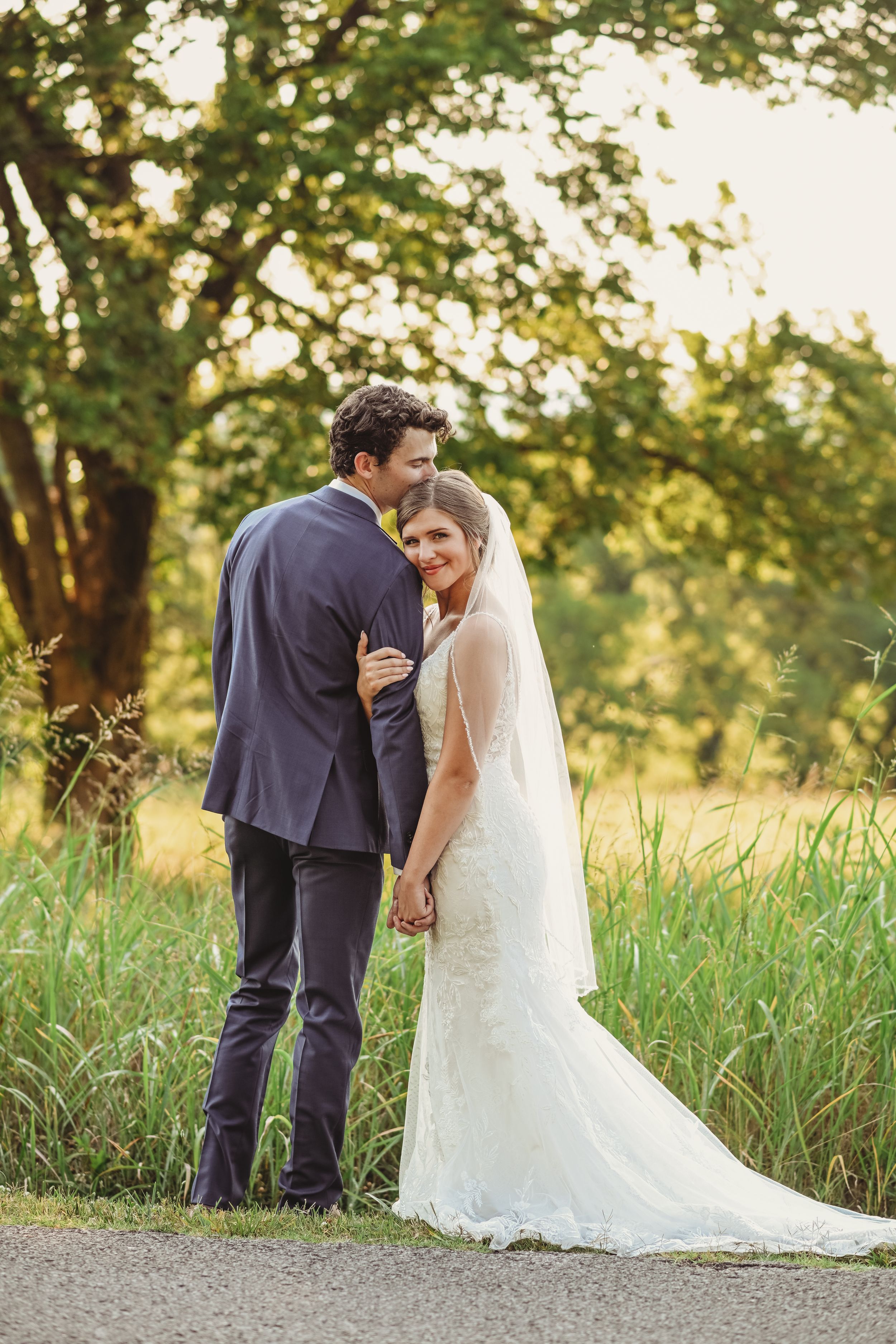 Newlyweds share intimate moment on path surrounded by tall summer grass.