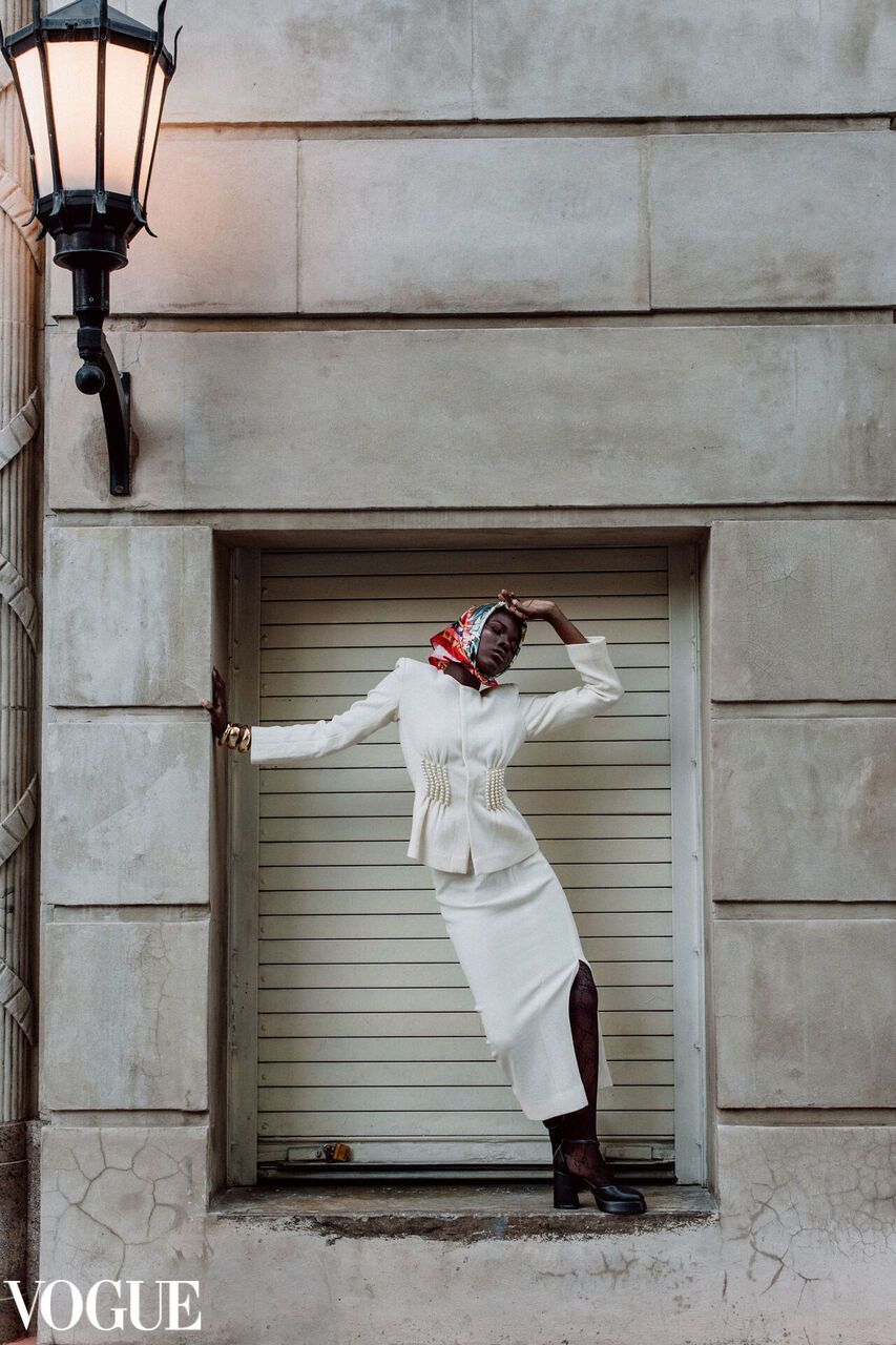 Fashion model strikes dramatic pose in white coat against industrial doorway with vintage lamp in moody Vogue photoshoot.