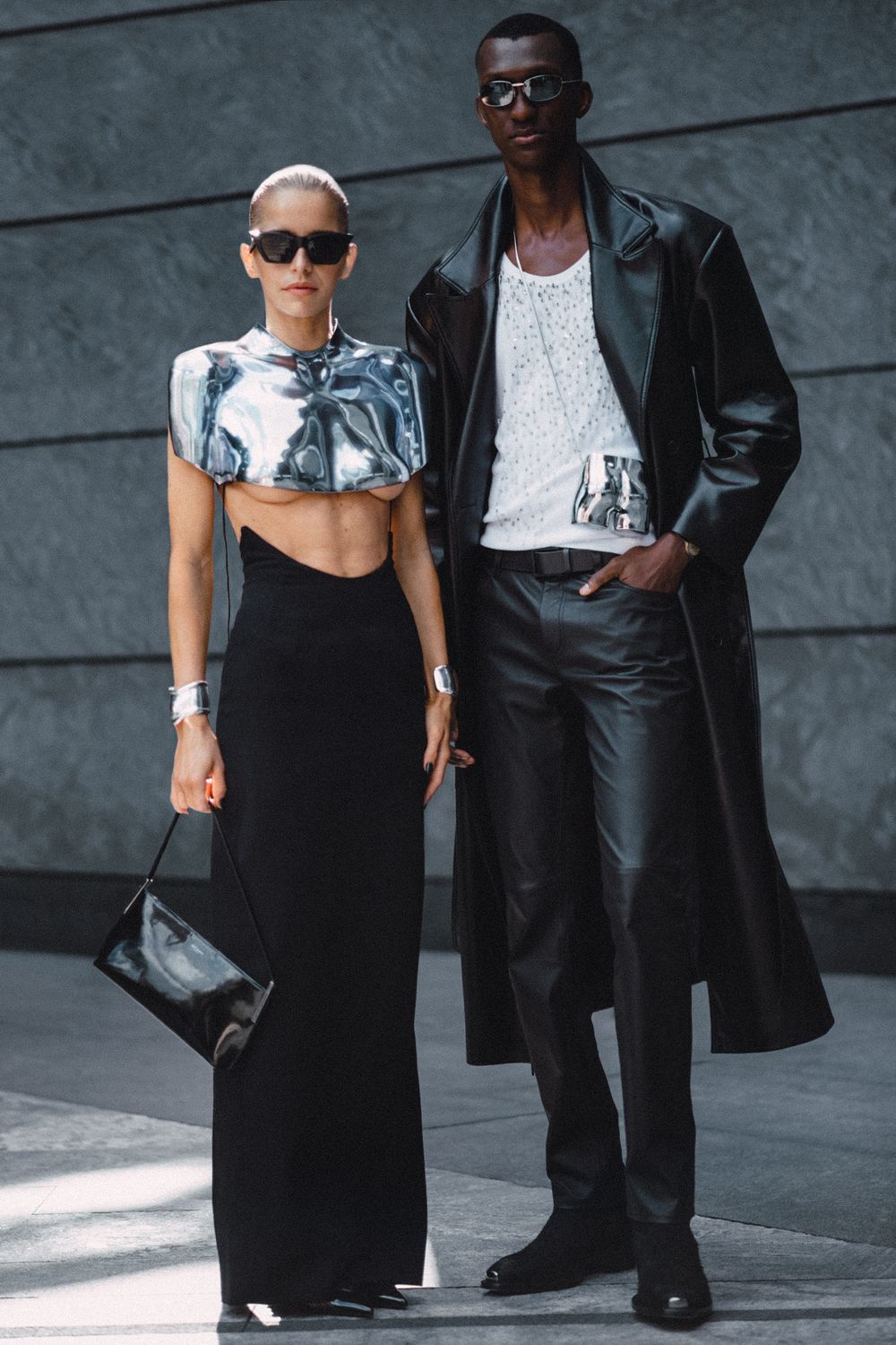 Fashion-forward couple in modern streetwear poses against urban wall wearing black and white ensembles.