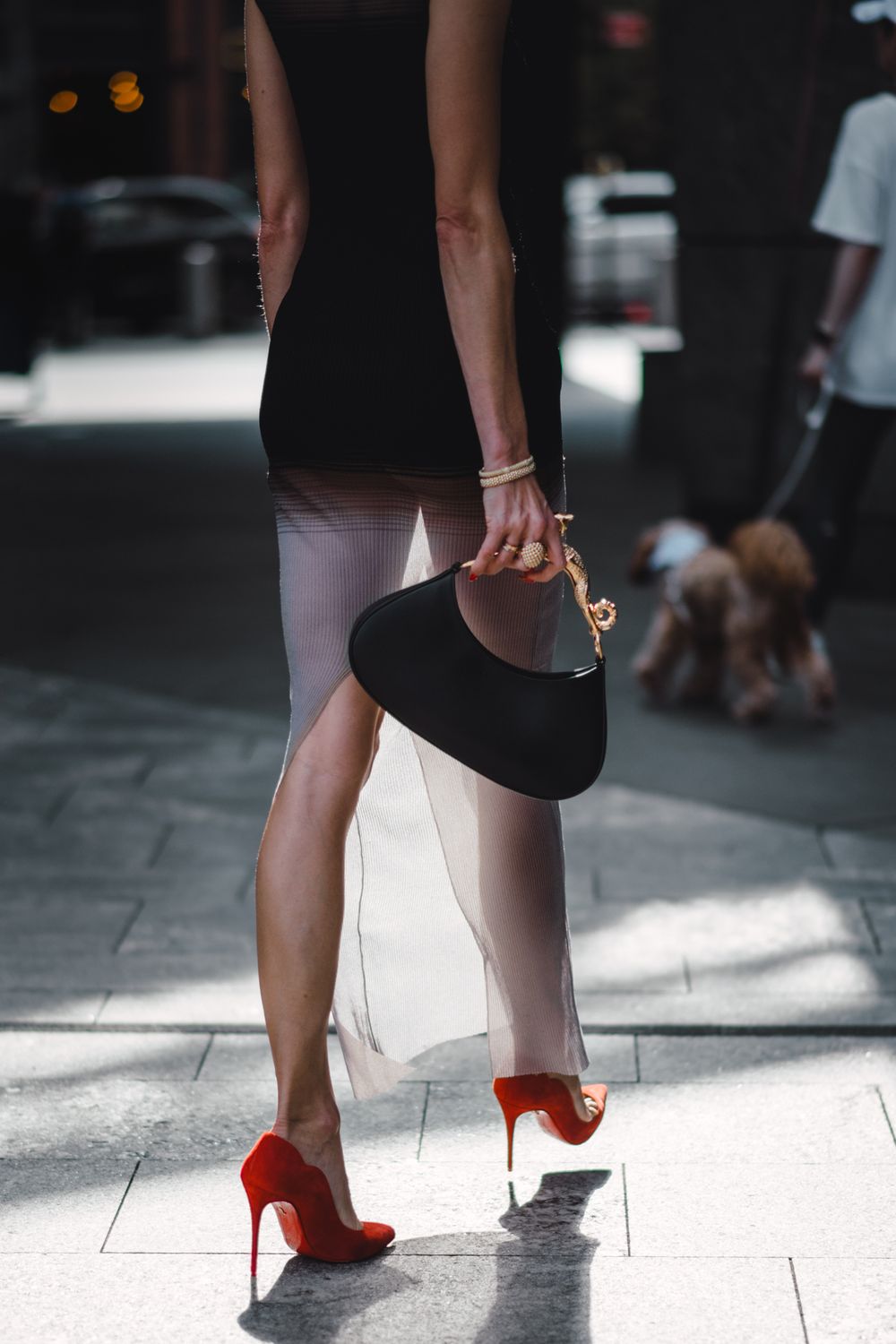 Close-up of legs in red stilettos and black dress holding designer handbag on city sidewalk.