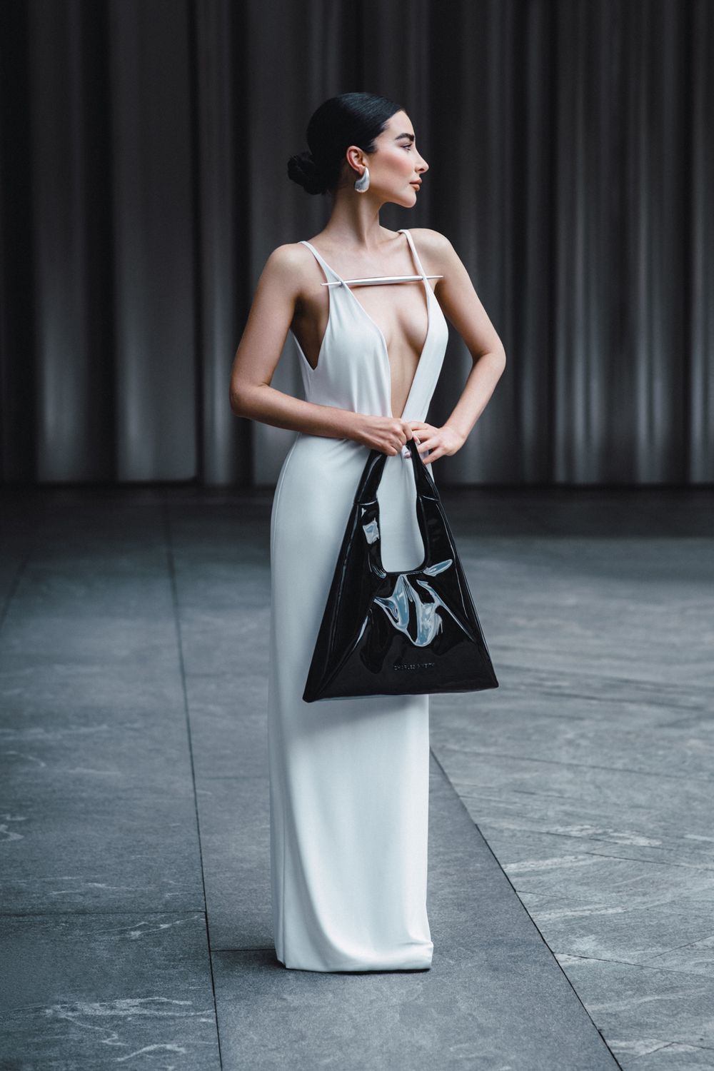 Elegant woman in white column dress poses with black patent leather handbag against dark curtained backdrop.
