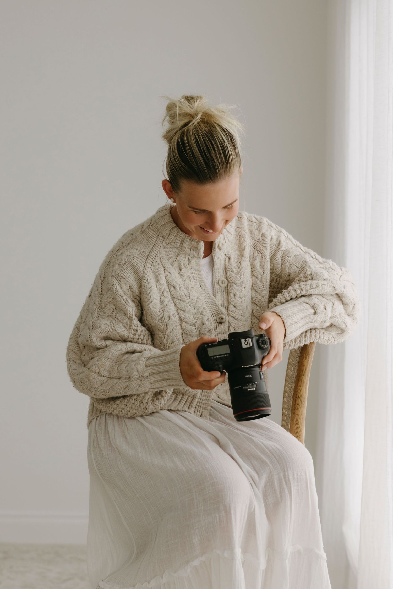 Female photographer in knit cardigan reviewing images on camera while seated in chair.