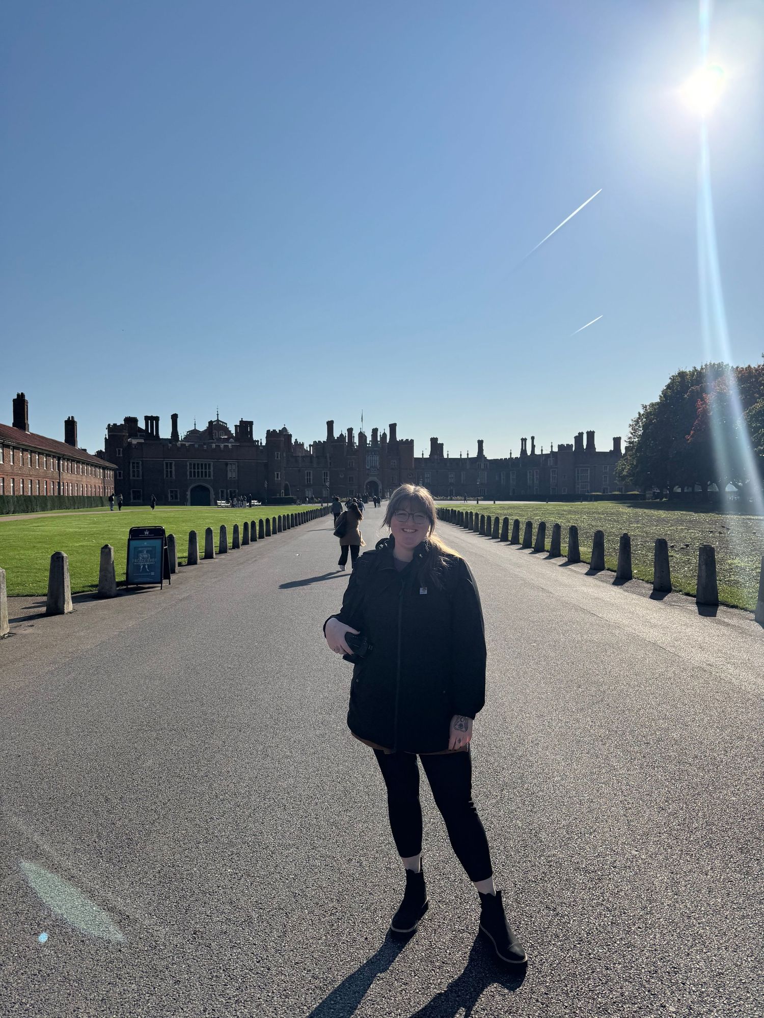 Person standing on long paved path with historic buildings and uniform columns Hampton Court on a sunny day.