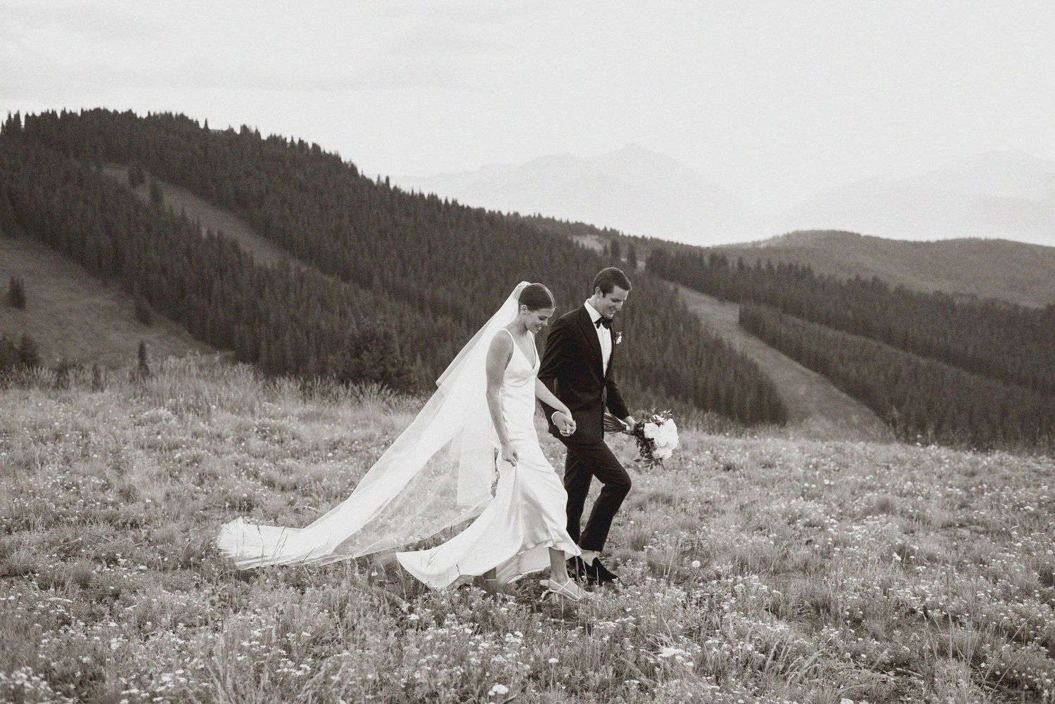 Black and white portrait of couple walking across mountain meadow in wedding attire.