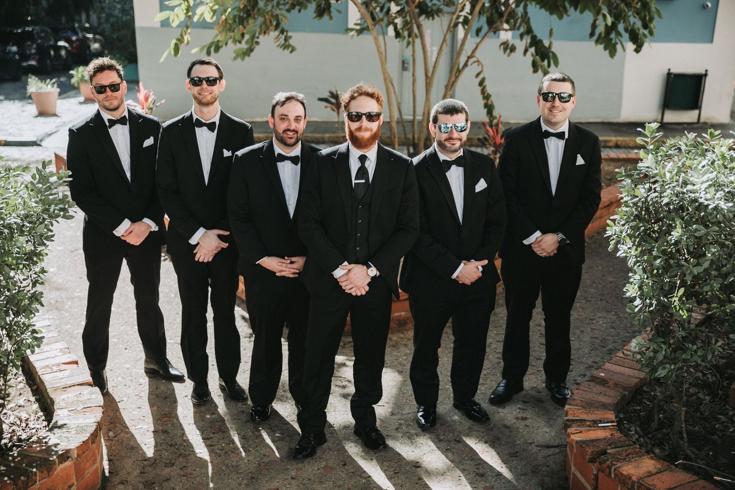 Wedding party of groomsmen in black suits and sunglasses pose together outdoors.