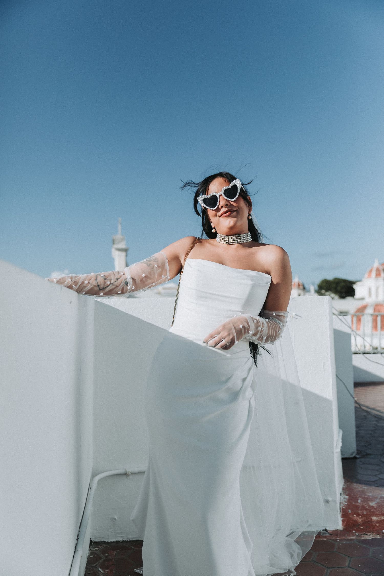 Joyful bride in sunglasses and white dress poses playfully on a rooftop.