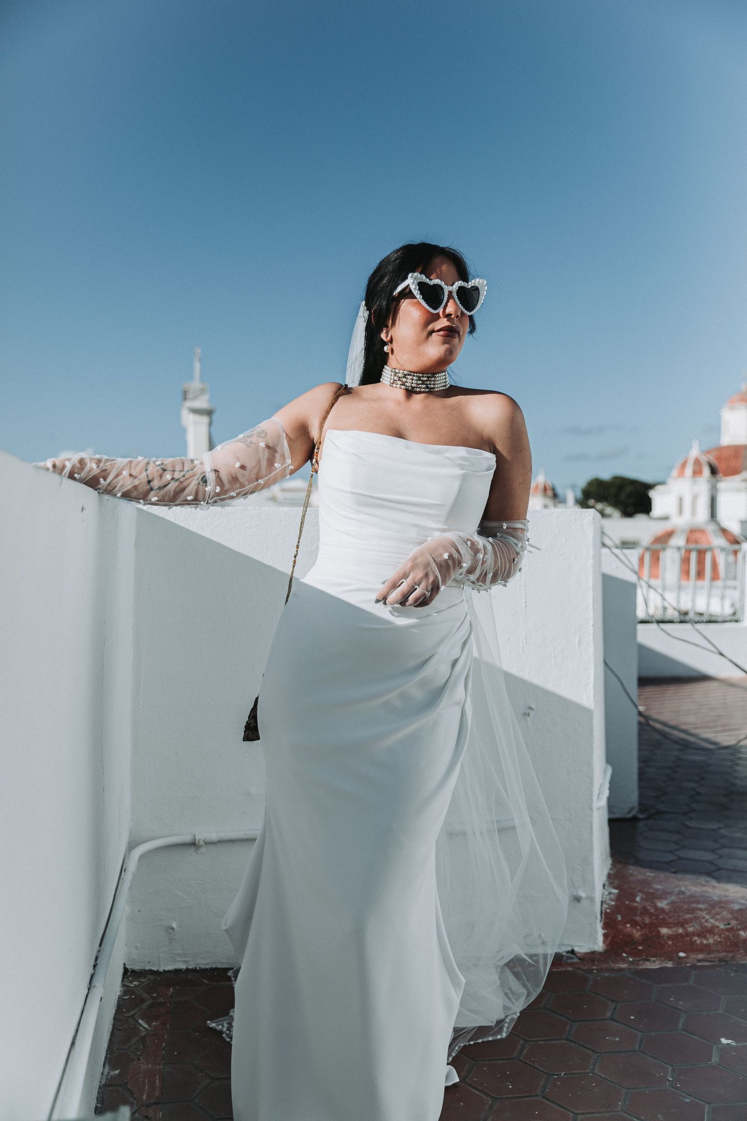 Stylish bride in sunglasses and white dress poses against bright blue sky on rooftop.