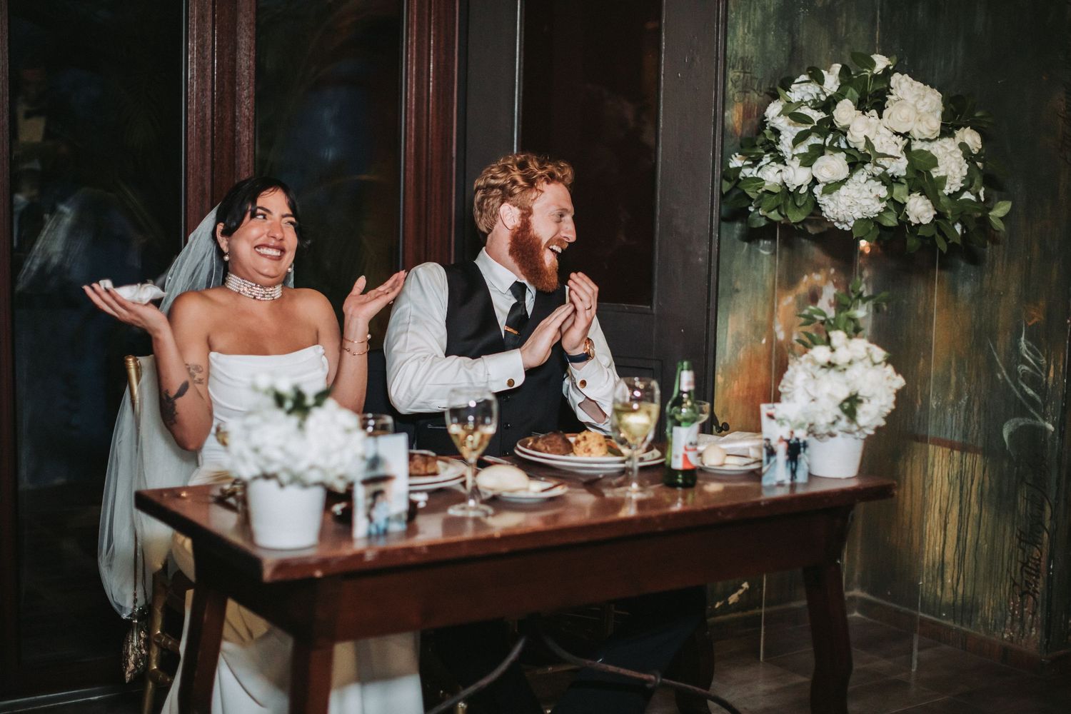 A bride and groom share a candid moment while eating at a rustic wooden table with floral decorations.