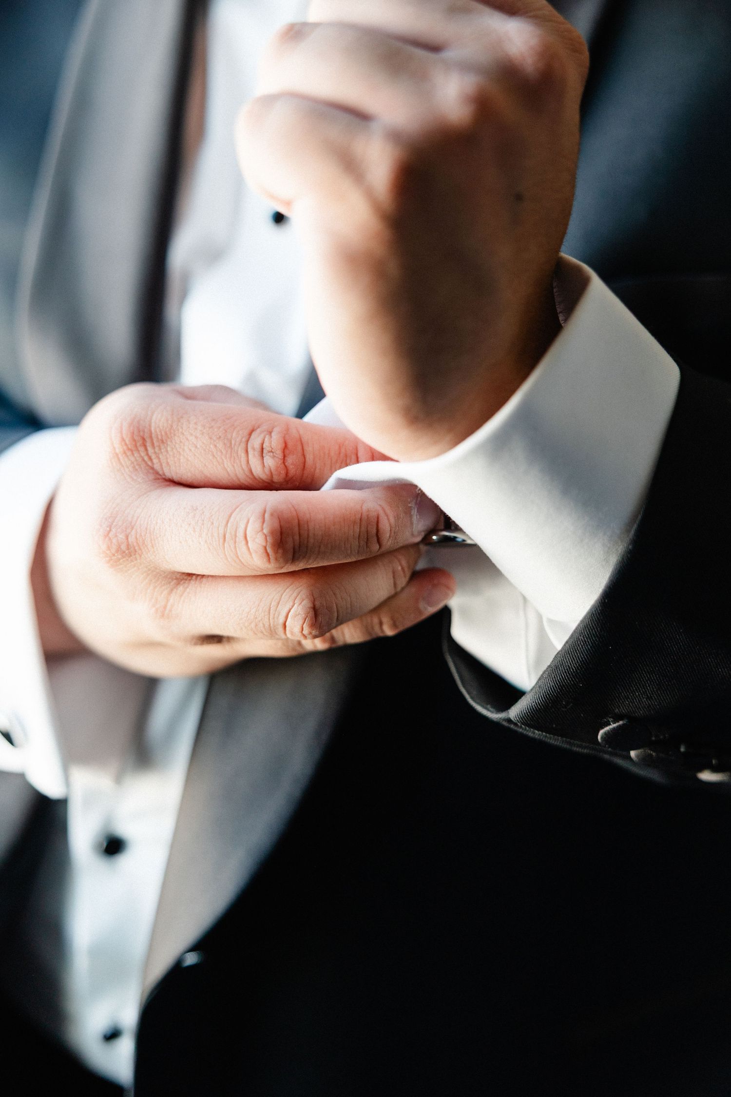 Man in a dark suit fastening a silver cufflink on a white dress shirt sleeve.