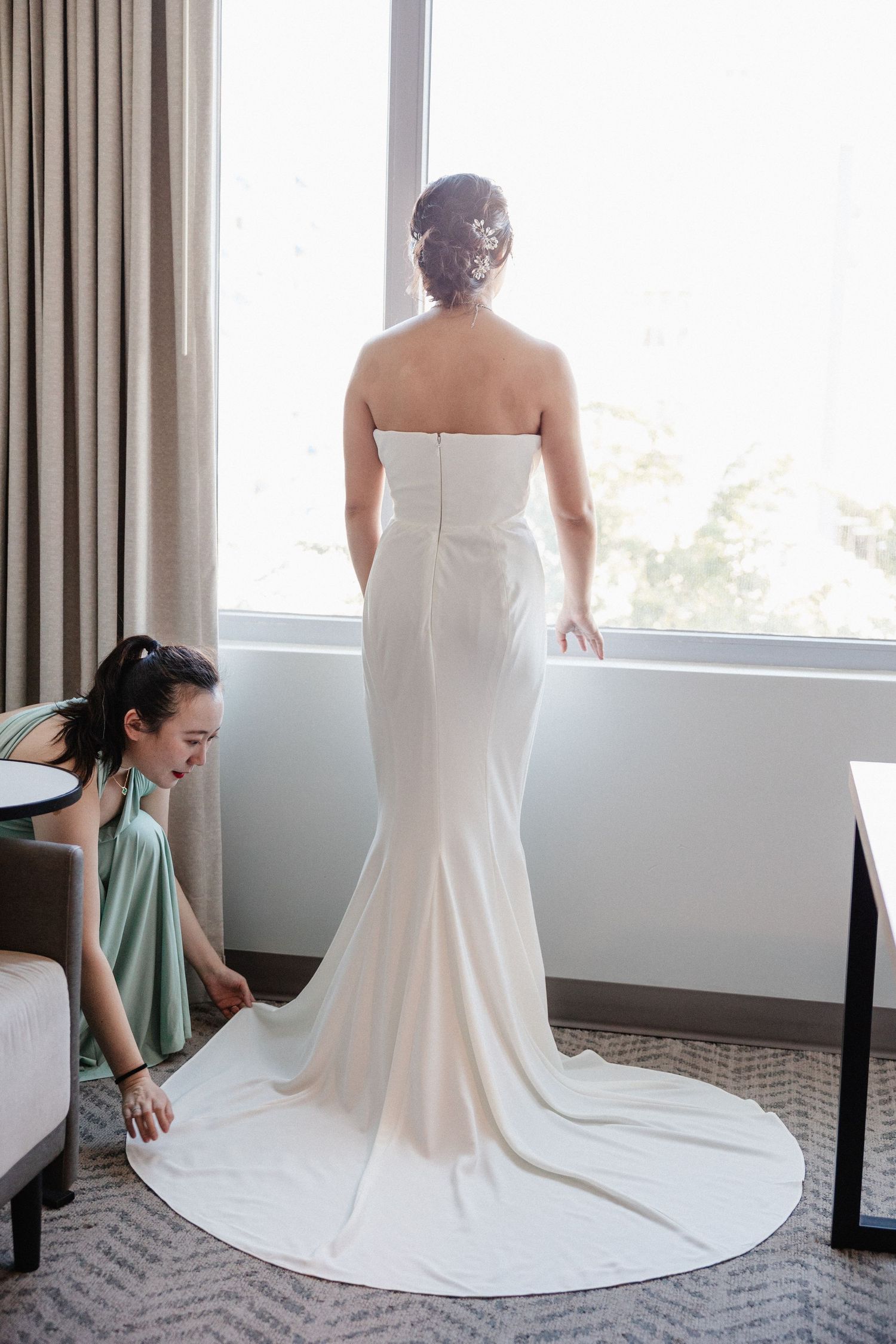 Bride in strapless white mermaid gown stands by window as bridesmaid in sage green dress arranges the train.